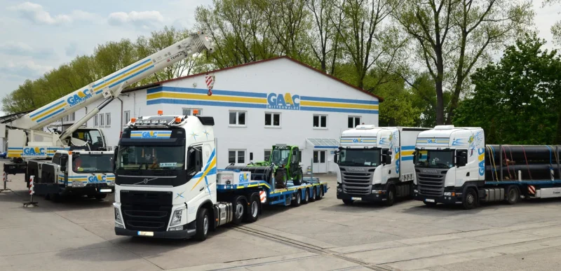GAAC Tieflader mit Baumaschine neben Autokran und Sattelzügen vor dem Firmengebäude
